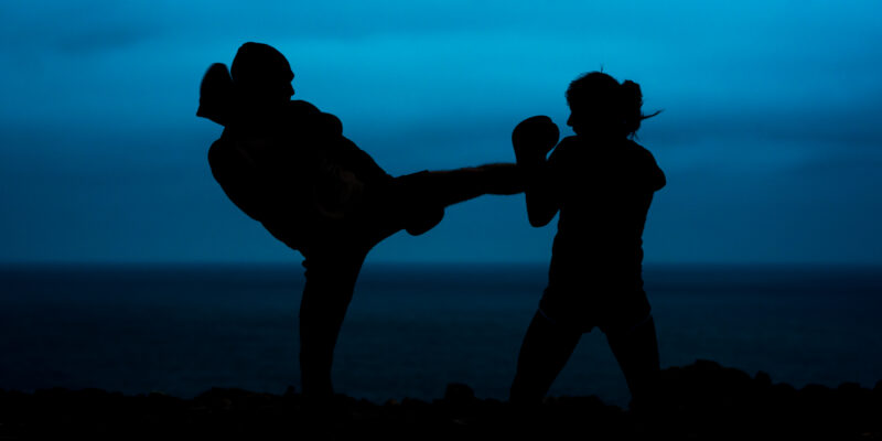 Silhouettes of anonymous fighters practicing kickboxing against sea and dark sky in evening in countryside